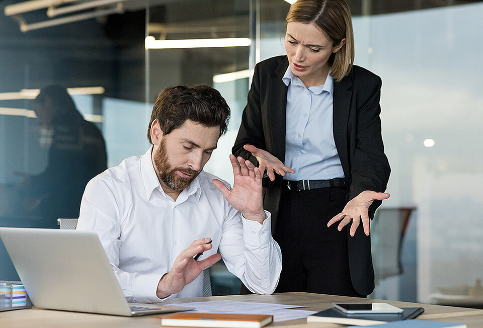 Eine Frau im Blazer diskutiert gestikulierend mit einem Mann am Schreibtisch, der abwehrend die Hände hebt. Symbolbild für den Streit um die Abnahme der Leistungsphase 9 im Architekturbüro.