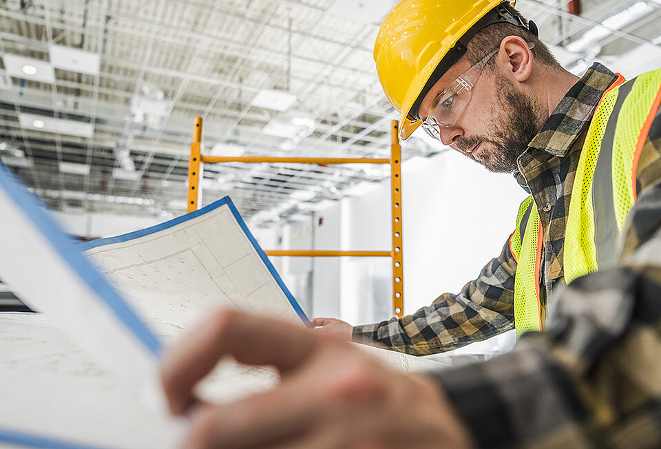 Ein Bauingenieur mit Helm und Schutzbrille prüft Ausführungspläne auf der Baustelle. Symbolbild für die konkludente Abnahme von Planungsleistungen durch Verwendung der Pläne bei der Bauvergabe.