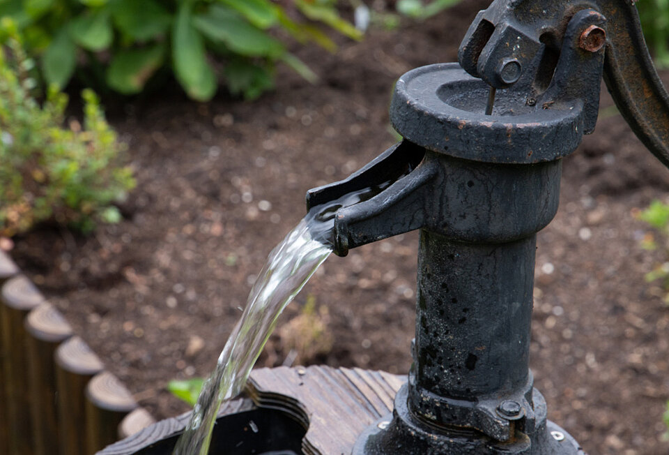Aus einer gusseisernen Schwengelpumpe im Garten fließt Wasser in ein Holzfass. Symbolbild für Rechtstipps zum Brunnenbau auf dem eigenen Grundstück.